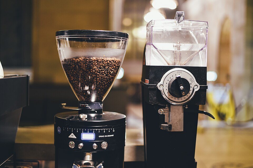 Two commercial grinders side by side on a café counter, one with a clear hopper full of roasted beans and a digital display, the other with a square hopper and a dial