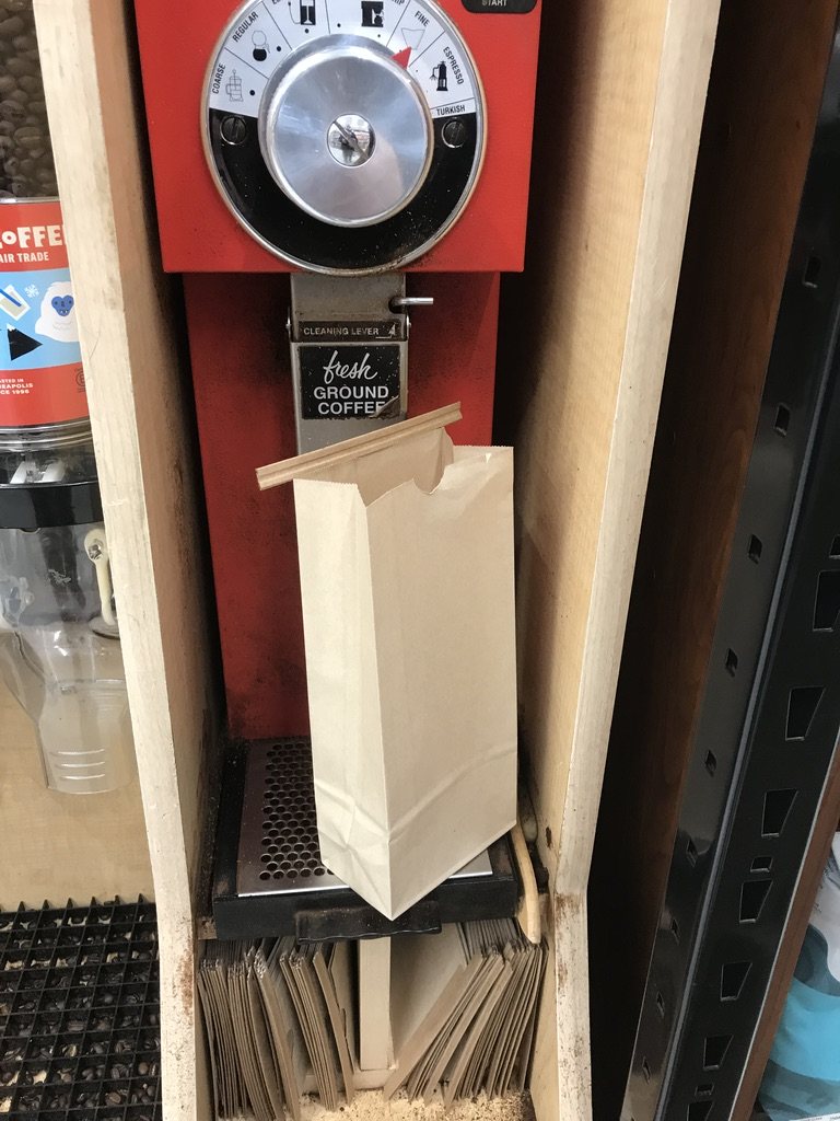 A red shop grinder with a paper coffee bag clipped under its spout, in a wooden stand in a store