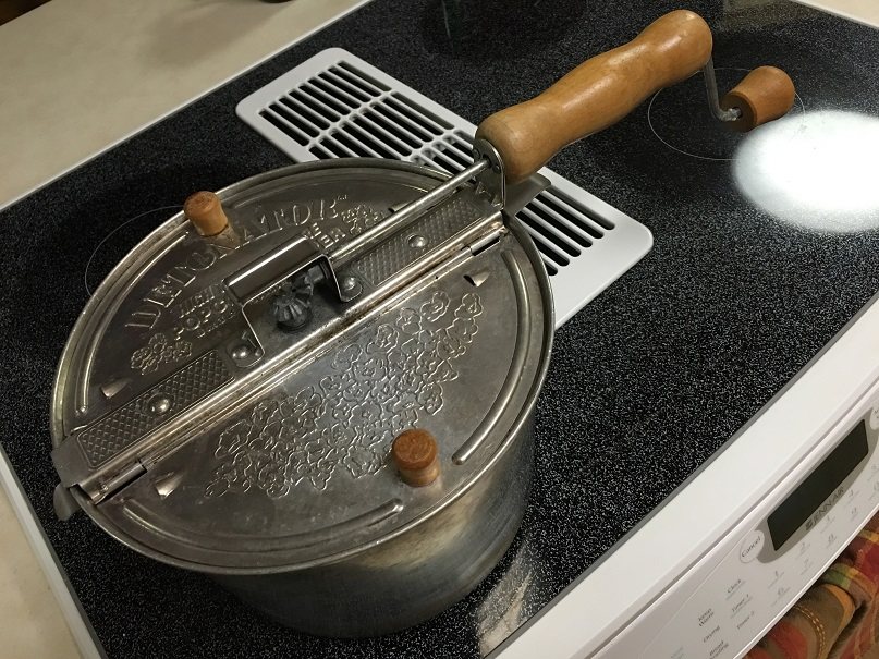 A stainless stovetop popcorn popper with a wooden handle and a crank in the lid, sitting on a glass-top range