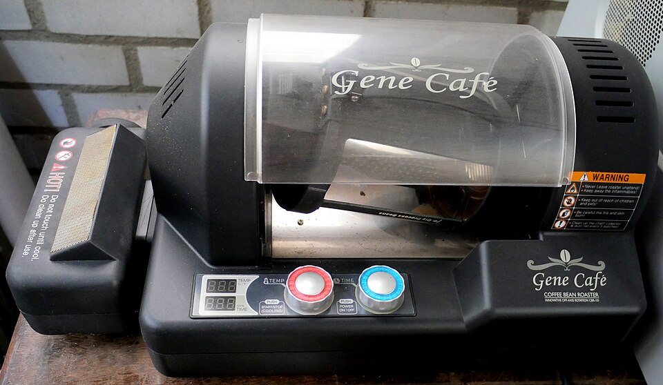 A Gene Café home coffee roaster: a black machine with a clear glass drum mounted at an angle and two dials on the front