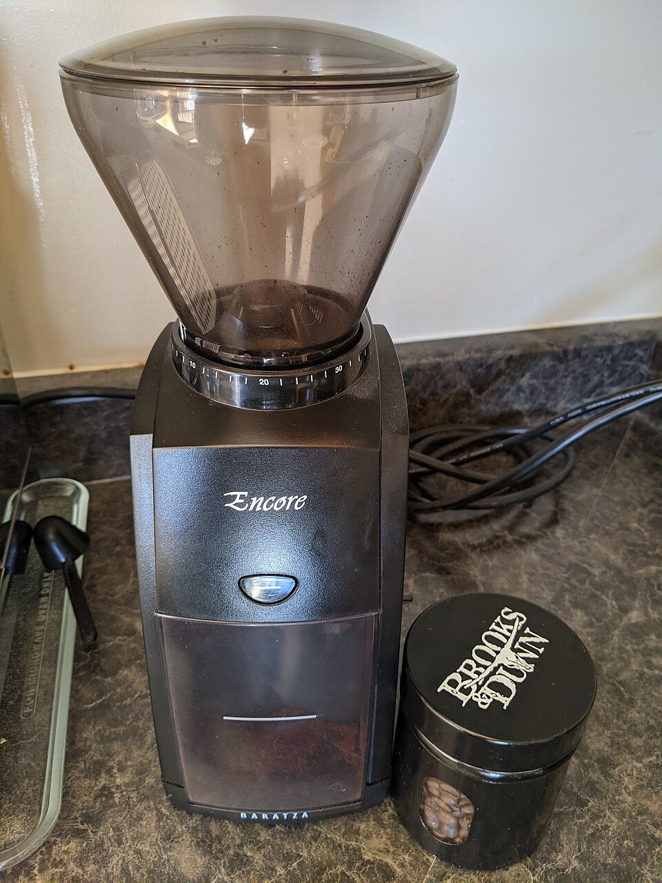 A Baratza Encore burr grinder on a kitchen counter, its clear hopper empty, with a small black tin of coffee beside it
