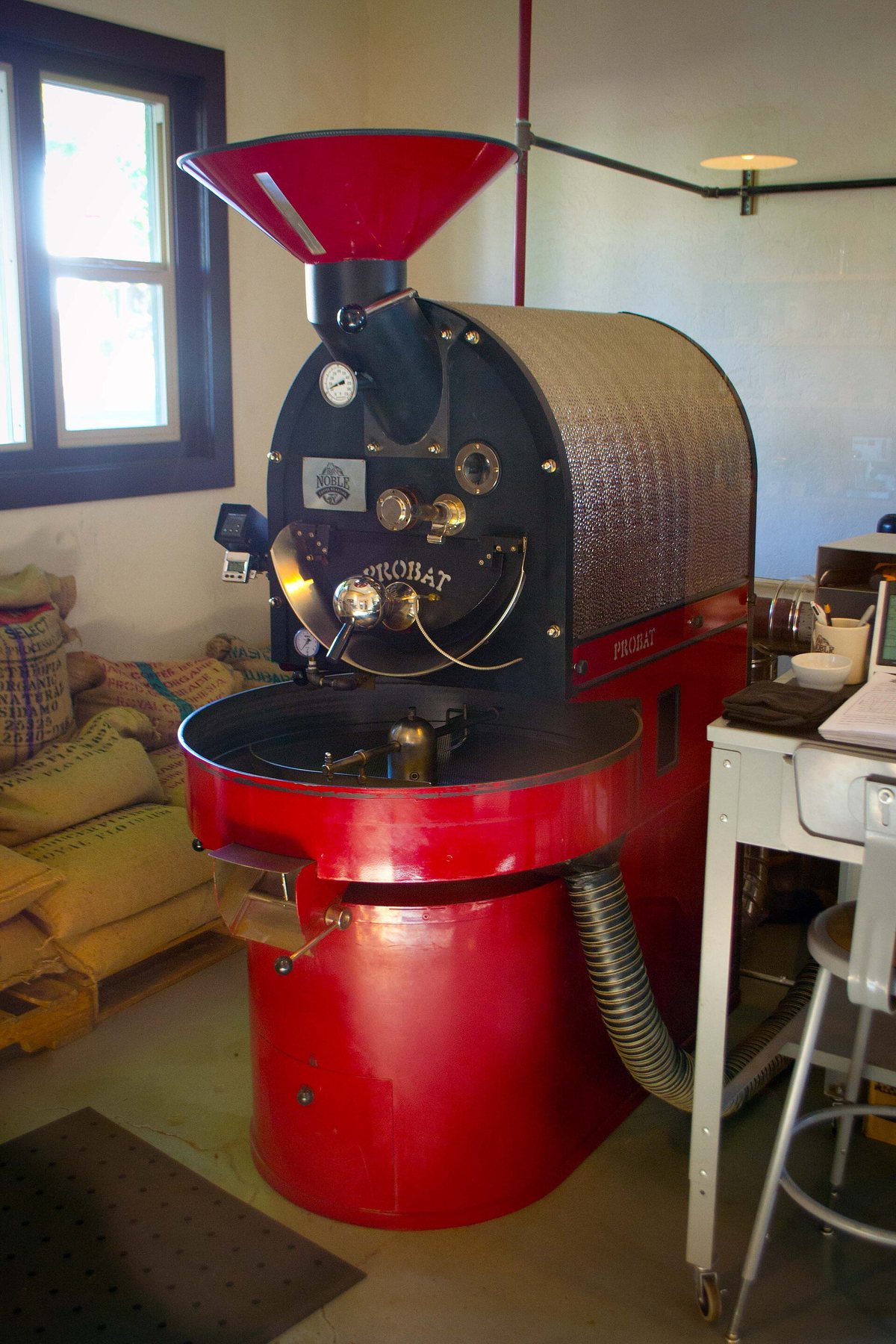 A shop drum roaster: hopper above, drum behind the faceplate, cooling tray with its stirring arm out front. Every rung of this page is a variation on these three parts