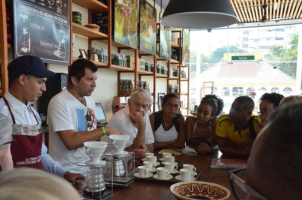 A barista in an apron explains a pour-over set-up to a group leaning in around a table of cups