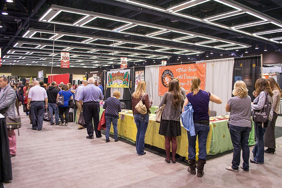 Visitors crowd around exhibitor tables on a convention-centre floor under numbered aisle banners