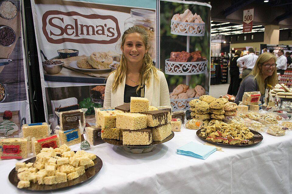 A vendor stands behind a table piled with crisped-rice bars and cookies at a trade-show booth