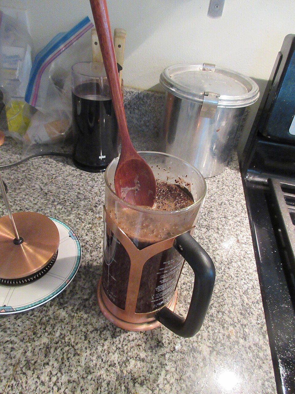 A French press with wet grounds floating at the top and a wooden spoon standing in it, the lid and plunger beside it on a counter