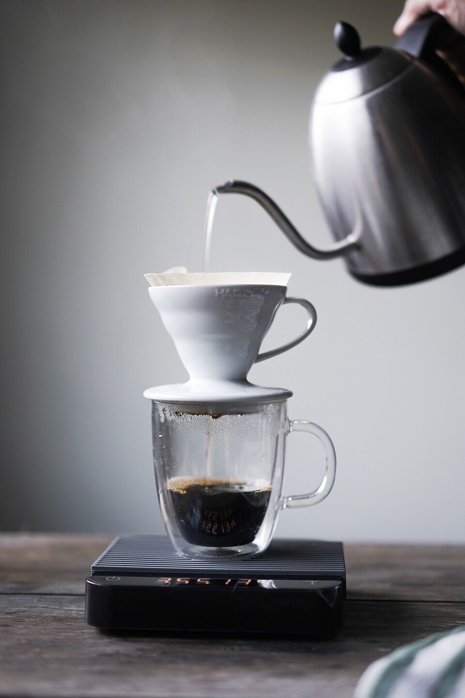 A white ceramic pour-over cone on a glass mug standing on a black digital scale, a gooseneck kettle pouring into it from above