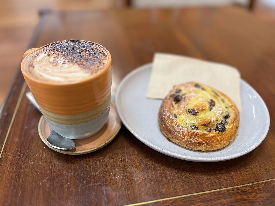 A cappuccino in a glass on a saucer beside a raisin pastry swirl on a plate