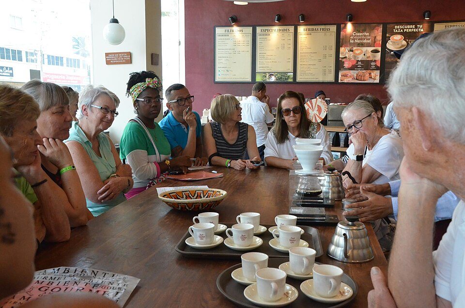 A dozen people seated around a long wooden table watching a pour-over being made, small cups set out in front of them