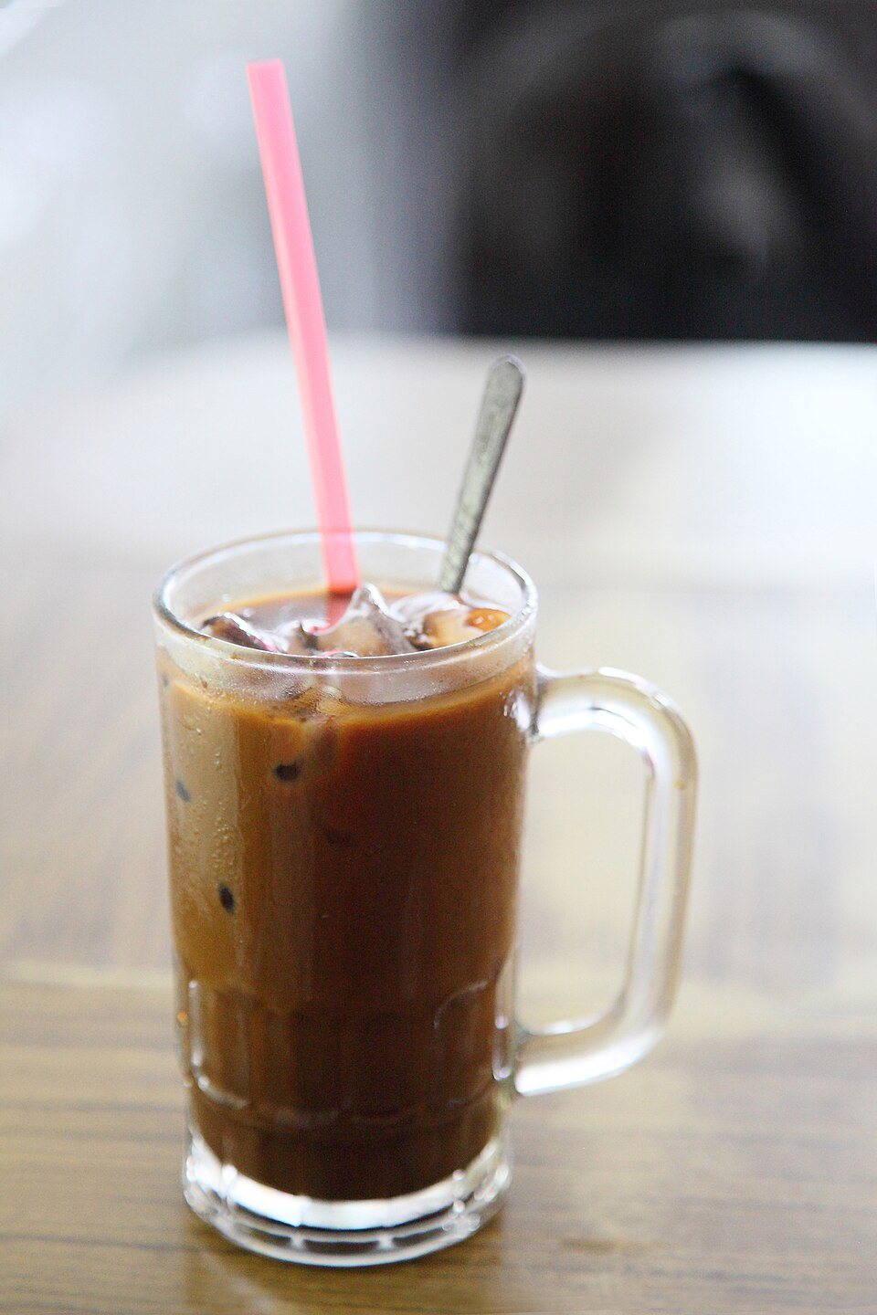 A glass mug of iced coffee with milk, a pink straw and a spoon standing in it
