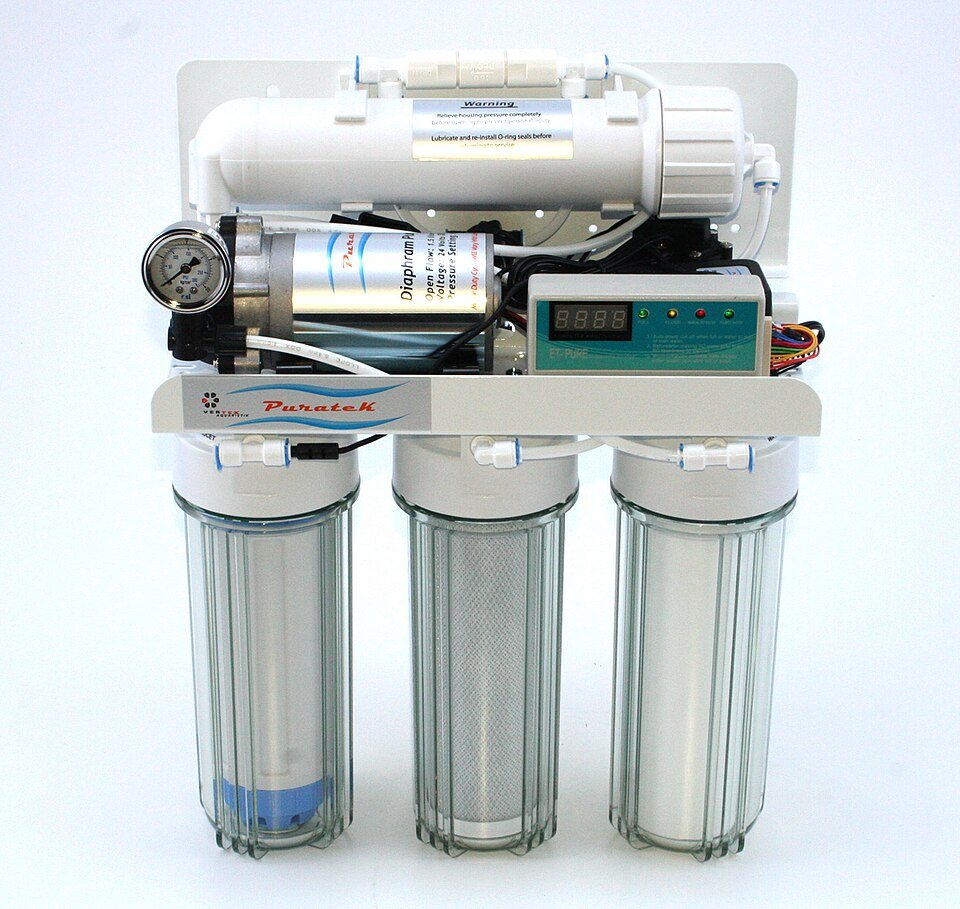 A small reverse-osmosis unit on a white backing plate: three clear filter housings in a row, a membrane tube across the top, a pump and a pressure gauge