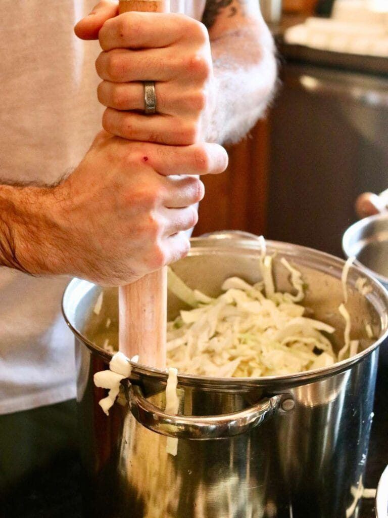 Pounding salted cabbage down with a wooden tamper. You are doing two jobs at once: rupturing cells to release brine, and compressing the mass under it
