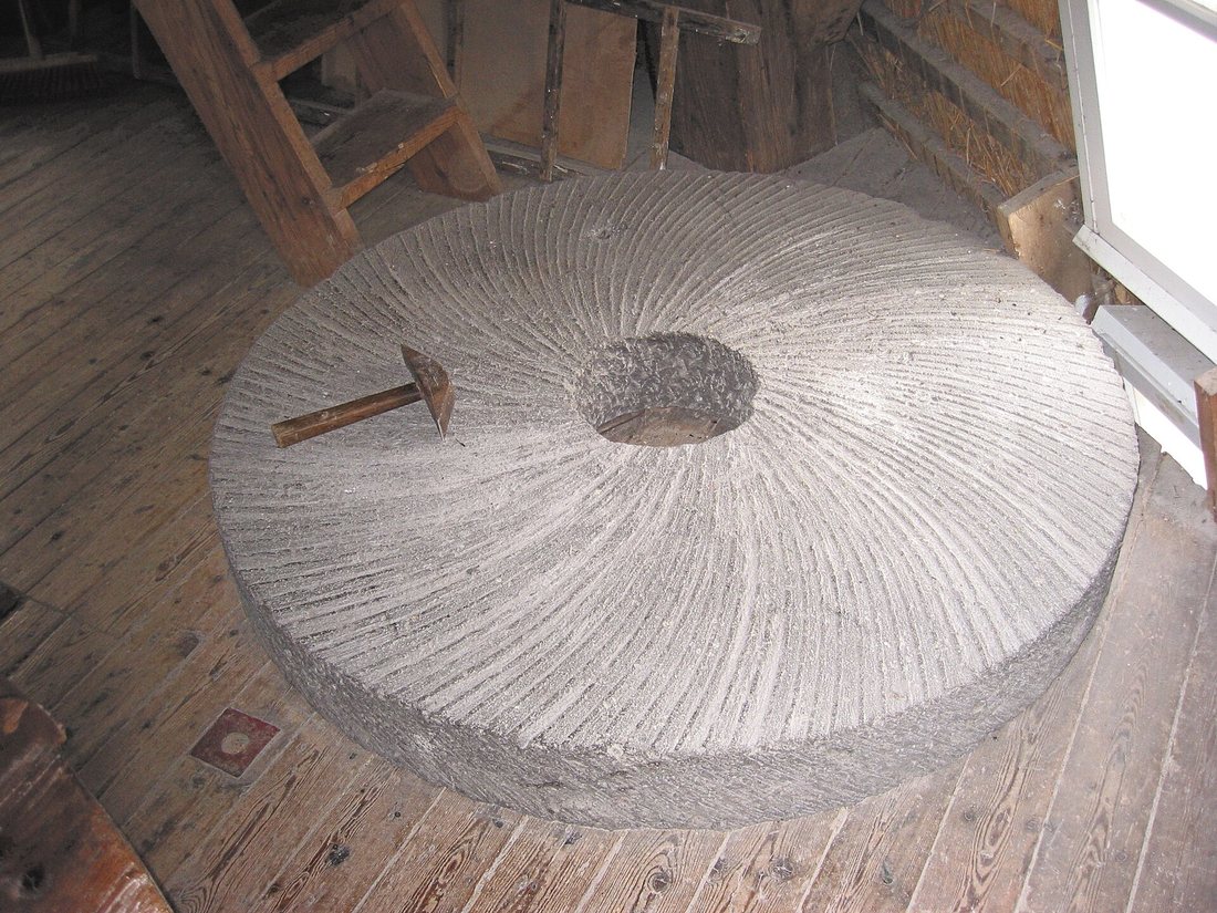 A millstone lying face-up on a wooden mill floor, its grinding face cut with fine curved furrows radiating from the central eye, a mill bill resting on it