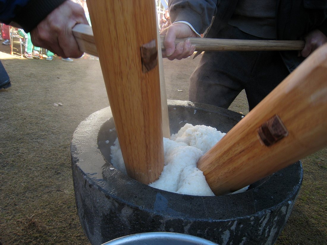 Two wooden mallets in a stone mortar full of steamed, partly pounded white glutinous rice