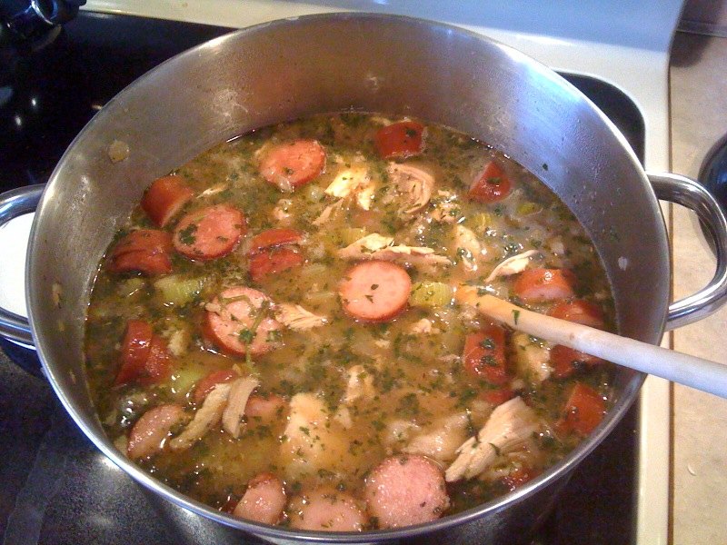 A pot of gumbo part-way through, sausage visible, being stirred