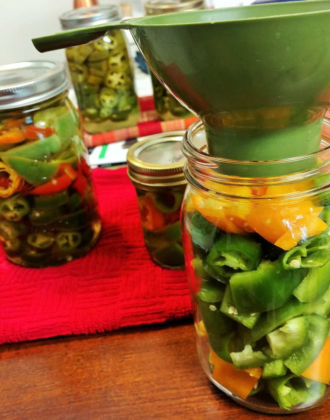 Jars of sliced peppers being filled through a canning funnel, more jars of pickled jalapeños behind