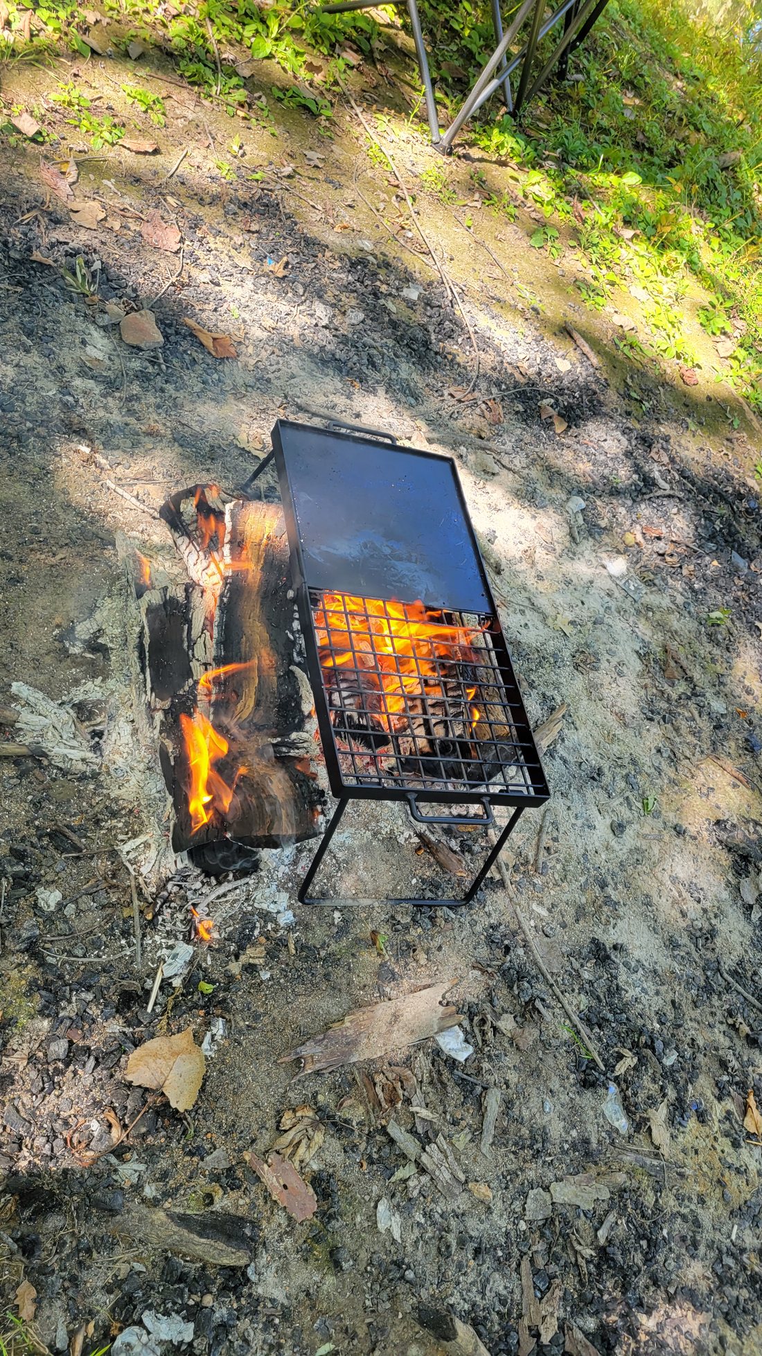 A folding camp grill, half solid griddle plate and half open grate, set over split logs burning in a fire ring