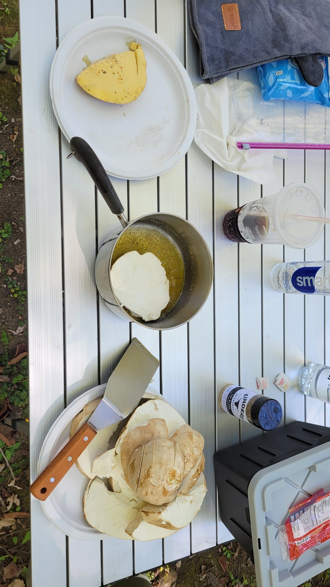 Slabs of a sliced giant puffball on a camp table, the cut faces uniform white, with one slab being dipped into a saucepan of melted butter and a shaker of salt-pepper-garlic seasoning nearby