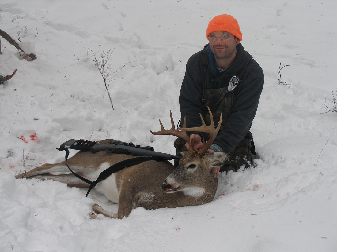 A hunter in blaze orange kneeling in snow beside a white-tailed buck