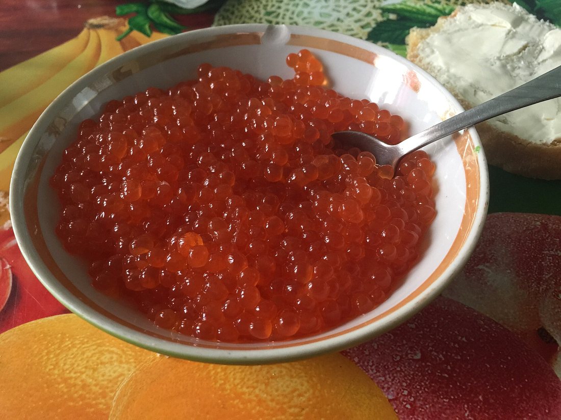 A bowl of bright orange salmon roe with a spoon, next to bread spread with cream cheese