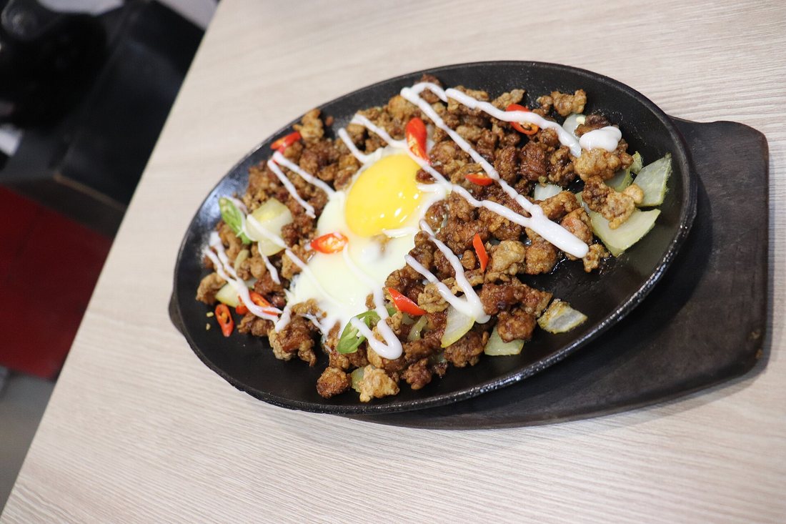 Pork sisig topped with an egg and mayonnaise on a black sizzling plate