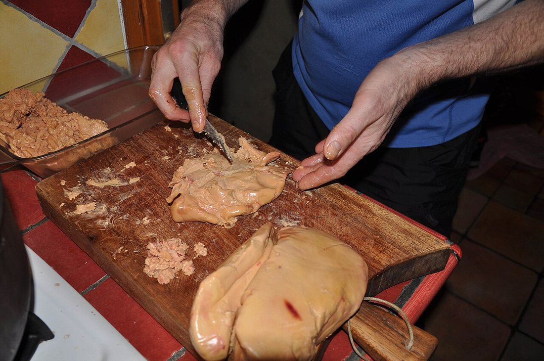Two hands deveining a lobe of raw foie gras on a wooden board, with a second lobe beside it