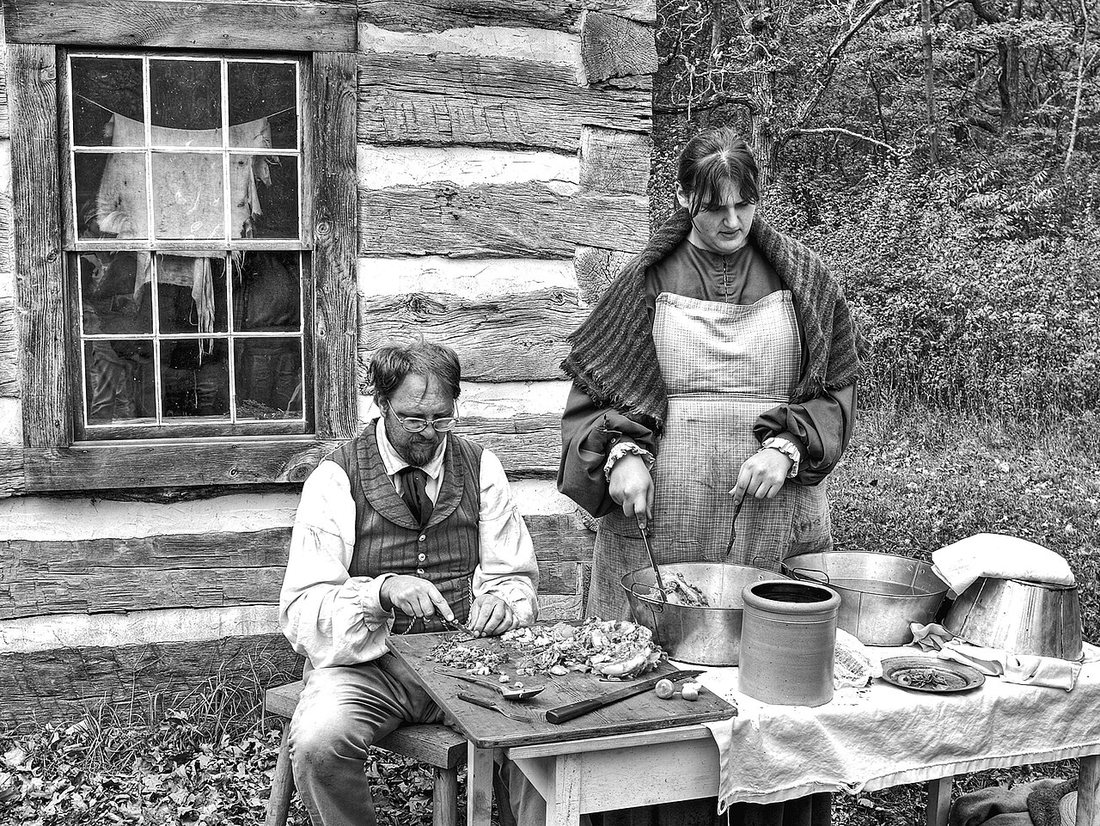 Black-and-white photograph of two re-enactors in period dress at an outdoor table beside a log house, preparing meat with crocks and pans