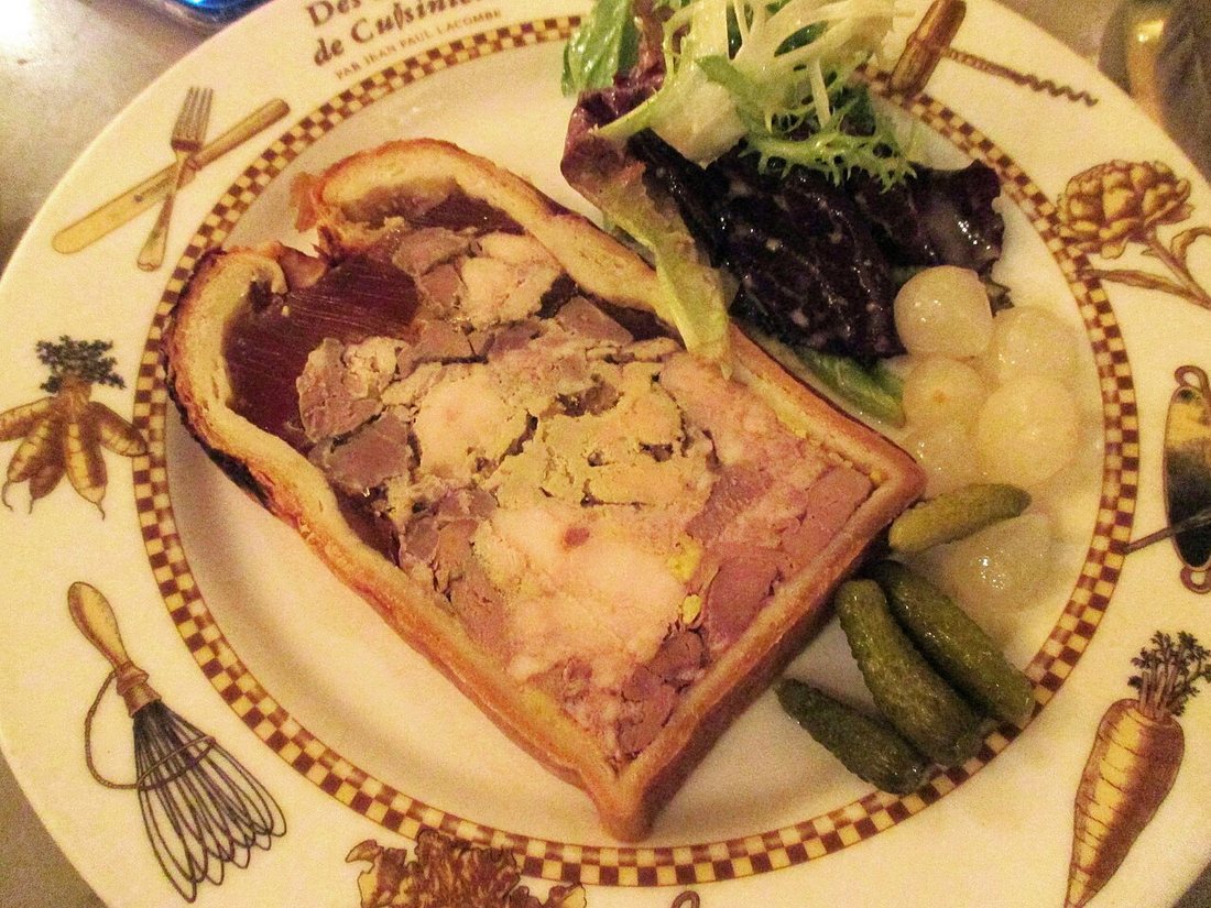 A slice of pâté en croûte on a decorated plate with salad, pickled onions and cornichons