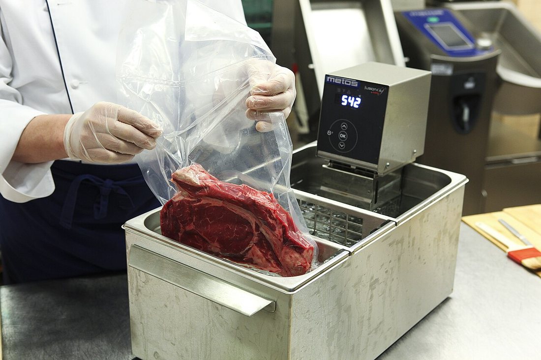 A cook in gloves lowering a vacuum bag of beef into a stainless-steel water bath with an immersion circulator reading 54.2