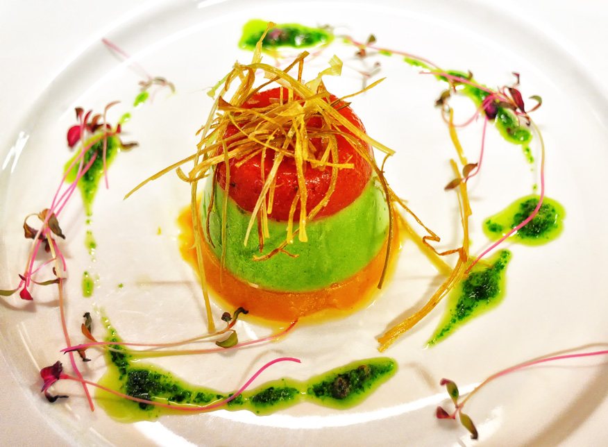 A small molded vegetable terrine in orange, green and red layers topped with crisp shreds, on a white plate with herb oil