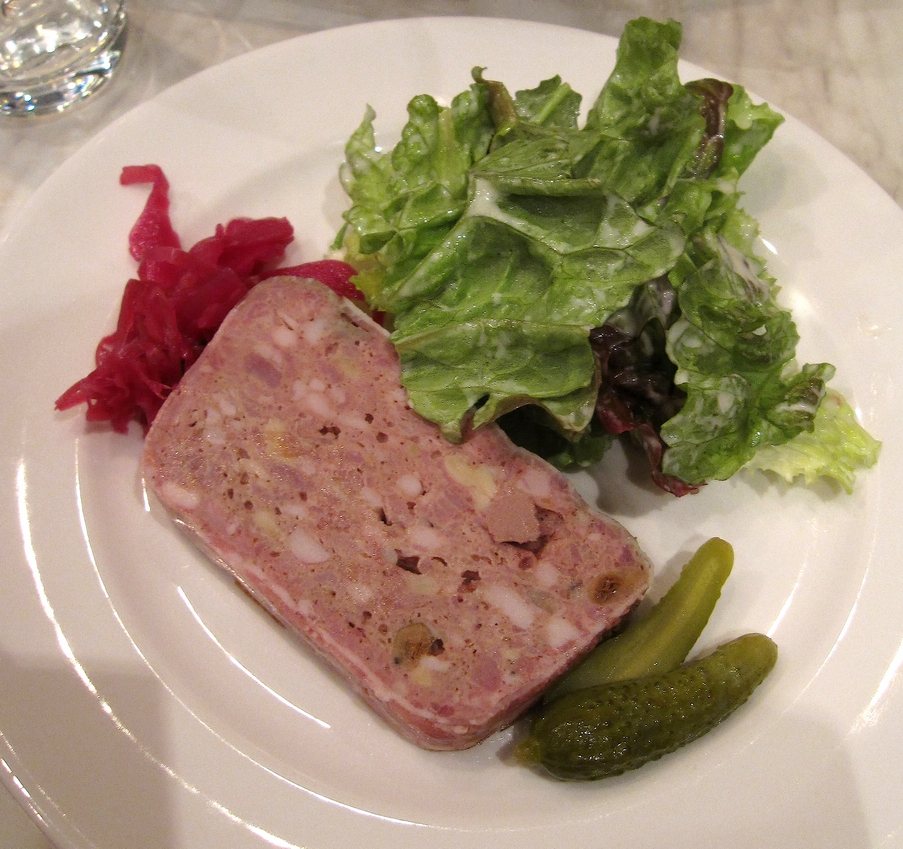 A slice of pink pâté de campagne with visible pieces of fat, served with lettuce, pickled red cabbage and cornichons