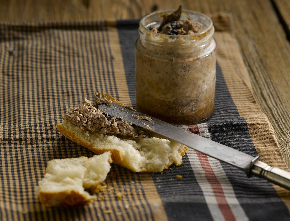A glass jar of coarse brown pâté on a striped cloth, with pâté spread on a torn piece of bread and a knife
