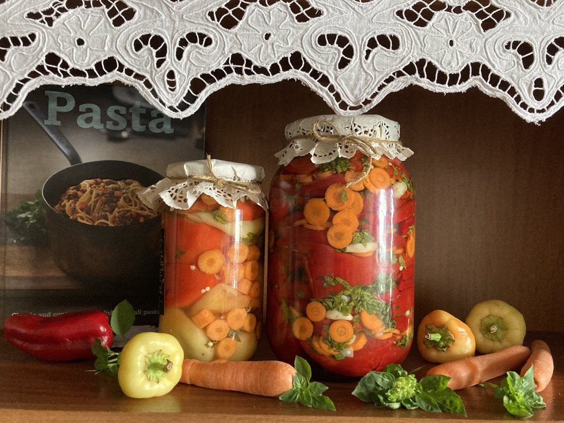 Two tall home-filled jars of red peppers, carrot slices, garlic and herbs, covered with cloth and twine, with fresh peppers and carrots arranged in front