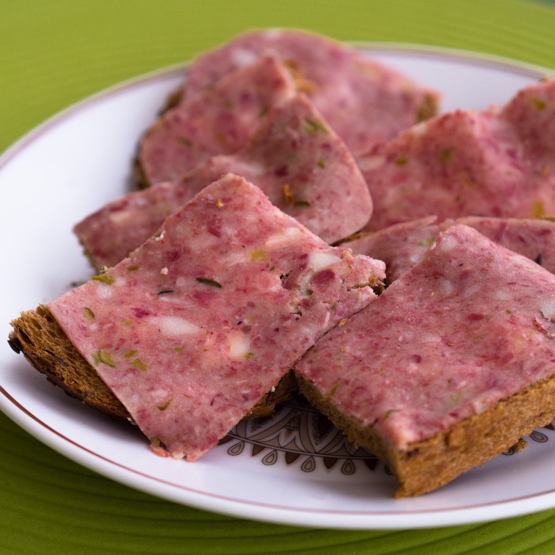 Slices of pale pink jellied meat, flecked with green herb, laid over dark rye bread on a plate