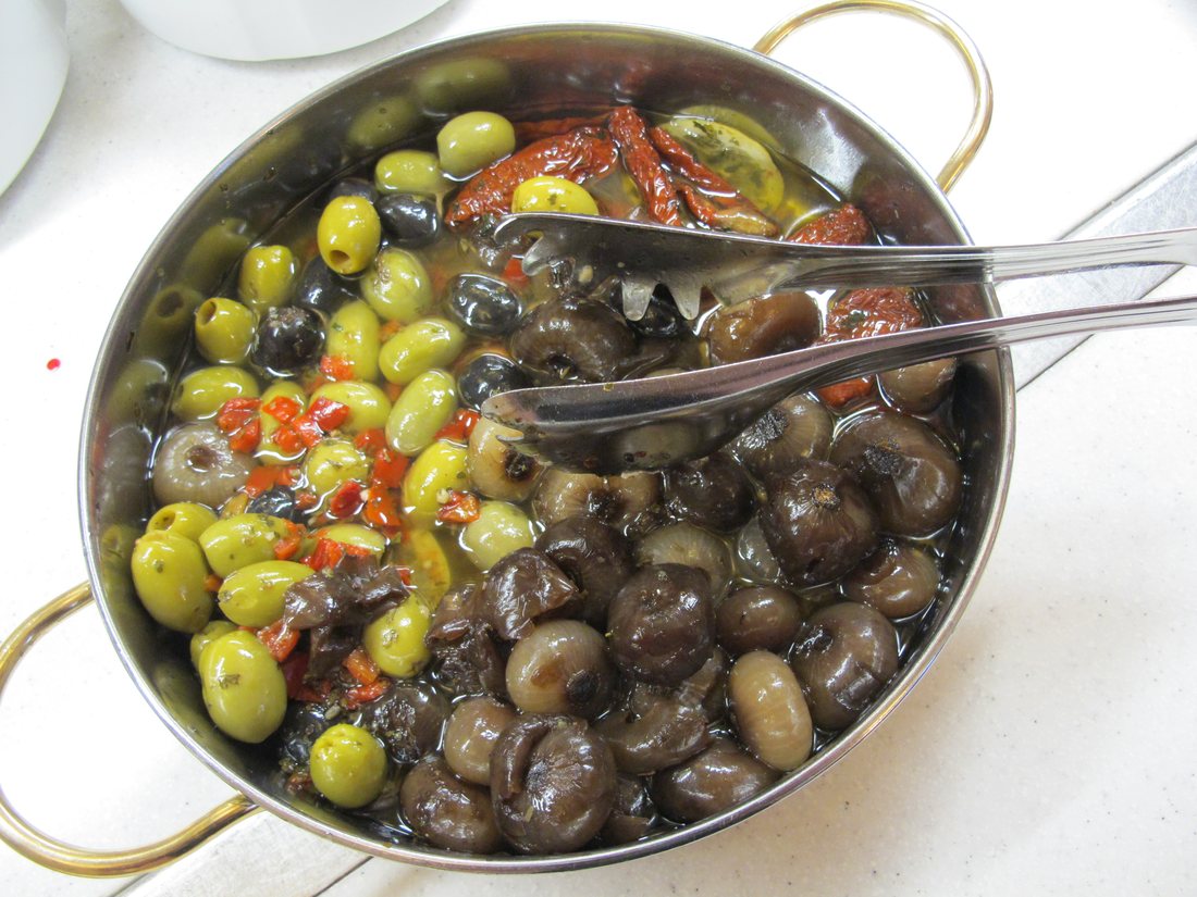 A metal serving dish holding green olives on one side and dark brown marinated mushrooms on the other, with two spoons and a little dried tomato between them