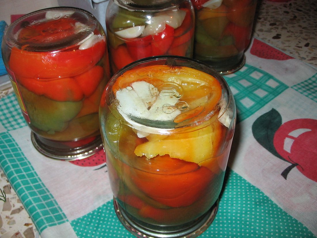Home-filled jars of red and orange peppers with whole garlic cloves, standing on a patterned oilcloth
