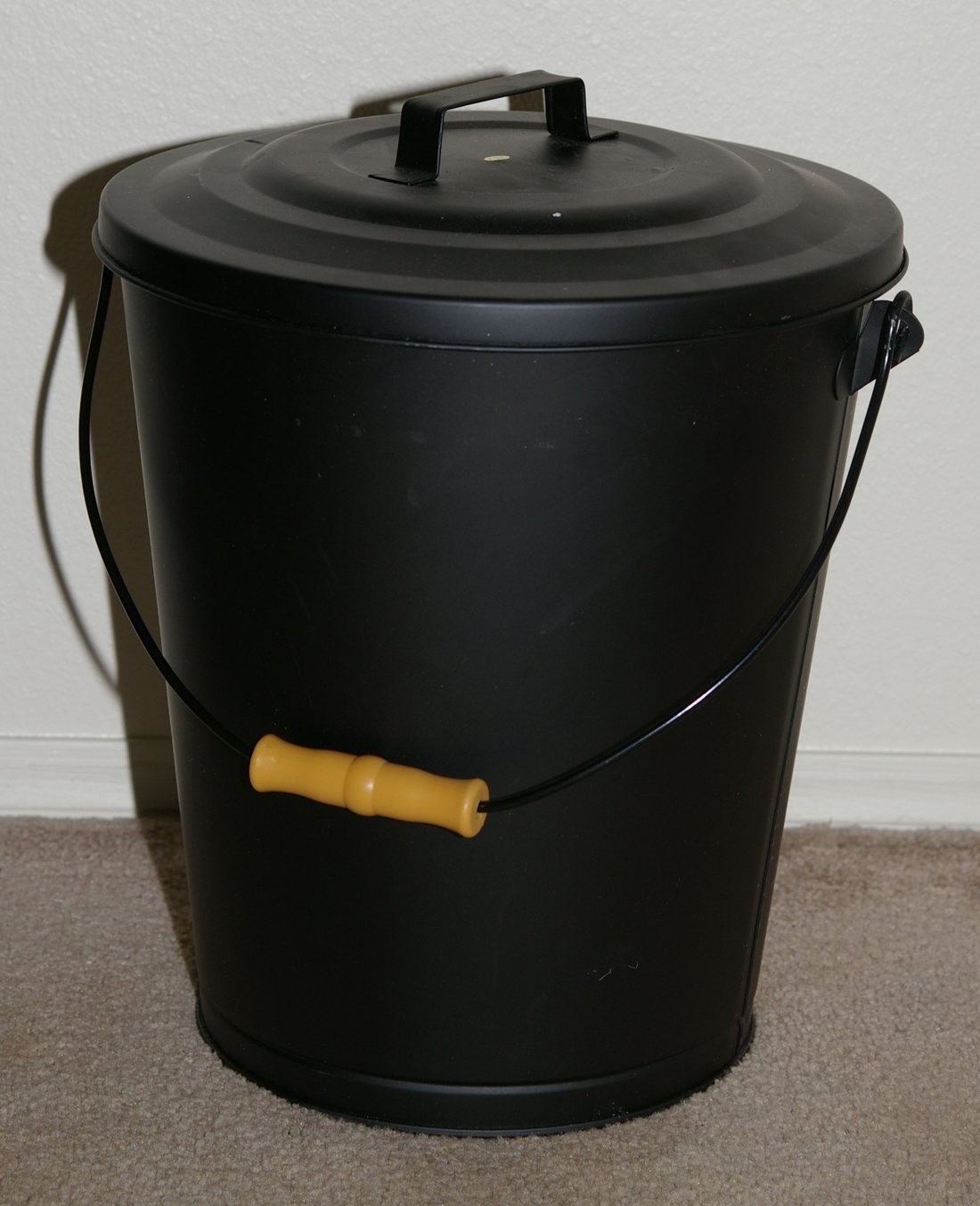 A black lidded metal bucket with a wooden handle standing against a wall