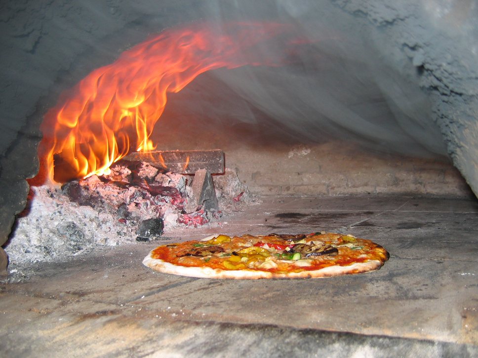 Inside a wood-fired oven: flames at one side curl up and across the dome while a pizza bakes on the floor beside the coals