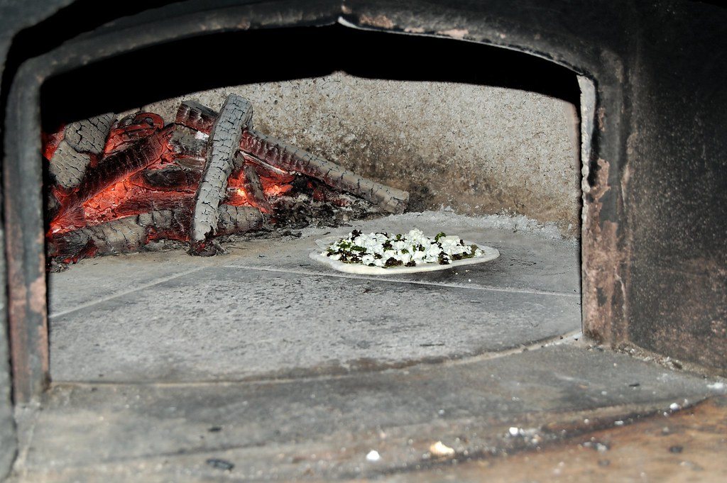 A flatbread topped with greens baking on the floor of a wood-fired oven, with embers and a log at the back