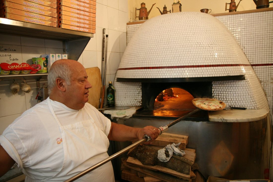 An older pizzaiolo in white holding a pizza on a long metal peel at the mouth of a white-tiled dome oven in Naples