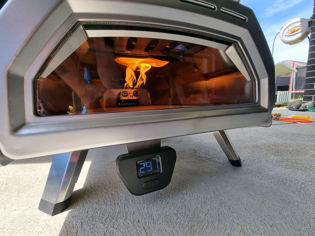 Close-up of an Ooni pizza oven with a glass door, flames from the rear fuel tray rolling across the roof, and a digital thermometer on the front