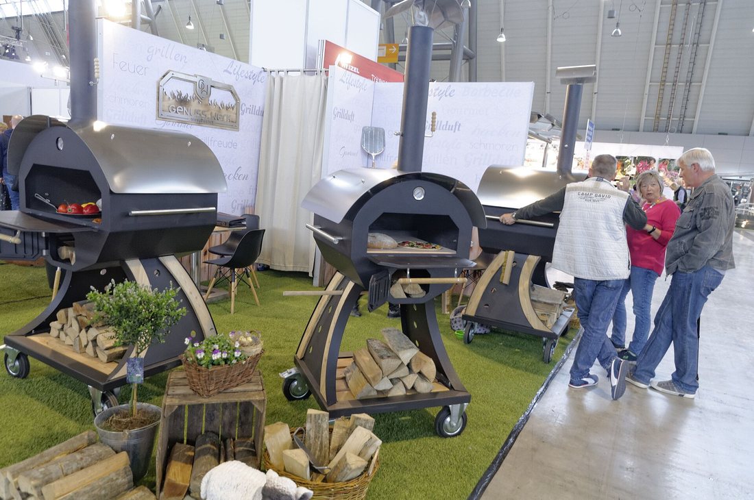 A trade-fair stand with a row of freestanding steel wood-fired ovens on wheeled carts, with logs stored underneath