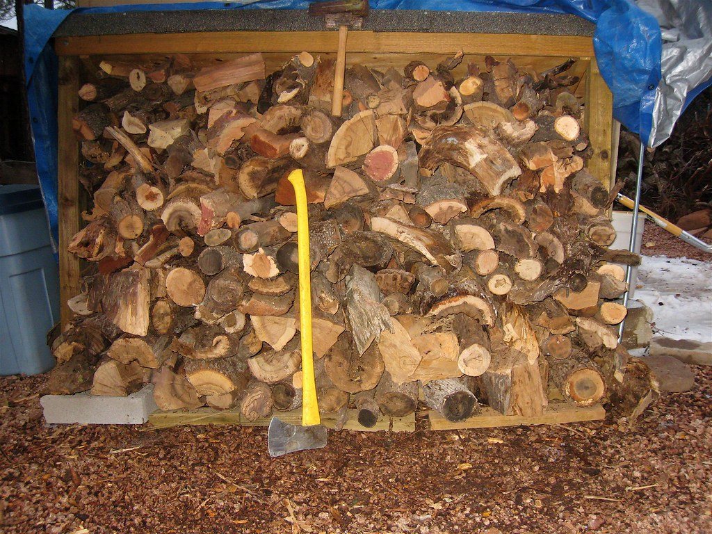 A bay of split firewood stacked under a tarp, with a yellow-handled axe leaning in front