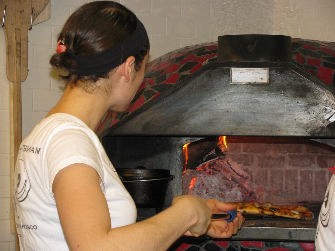 A cook using a small round turning peel to turn a pizza inside a tiled dome wood oven