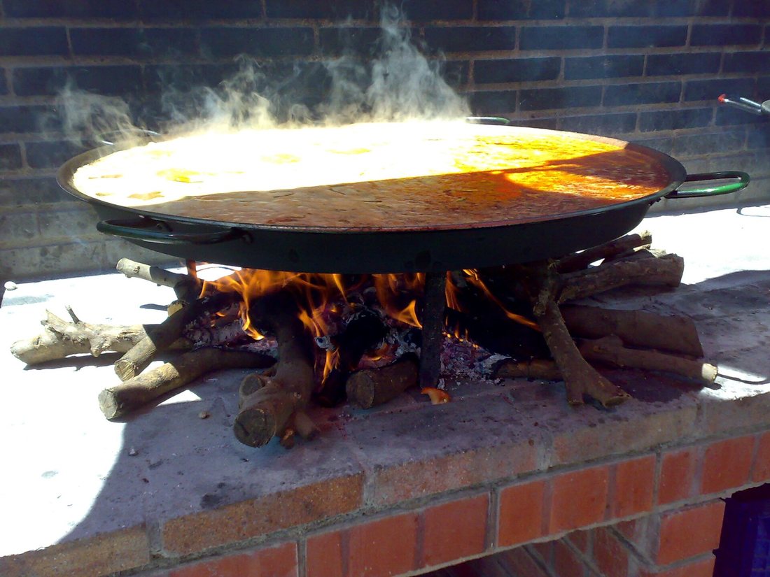 A paella pan over a wood fire on a brick outdoor hearth in Valencia, steam rising from the rice