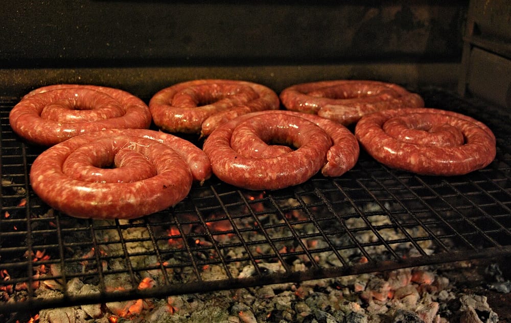 A large spiral of boerewors over an open wood fire, held flat by a grid, the coarse grind visible through the taut casing