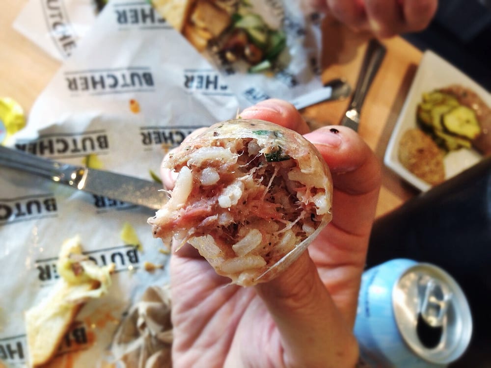 A Cajun boudin link held up at the butcher, the cut end showing its pale rice-and-pork filling — more rice than meat by volume