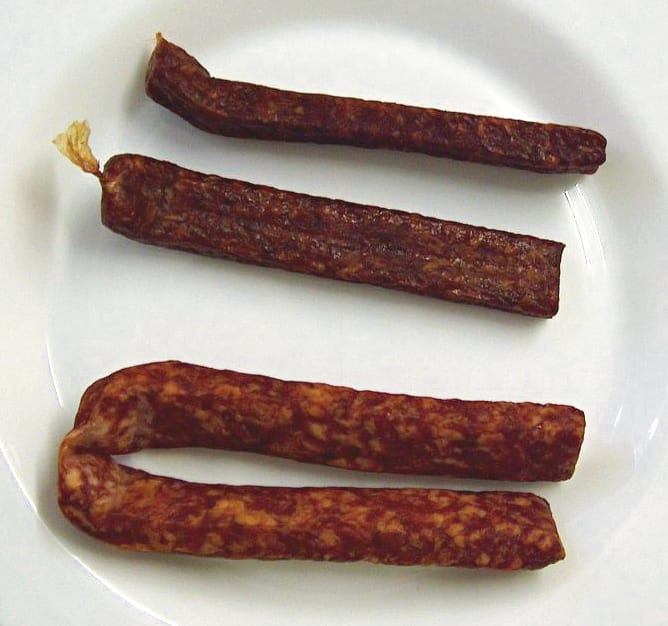 A pair of dark rectangular landjaeger still joined at one end, the flat pressed faces and squared edges obvious against a round link beside them