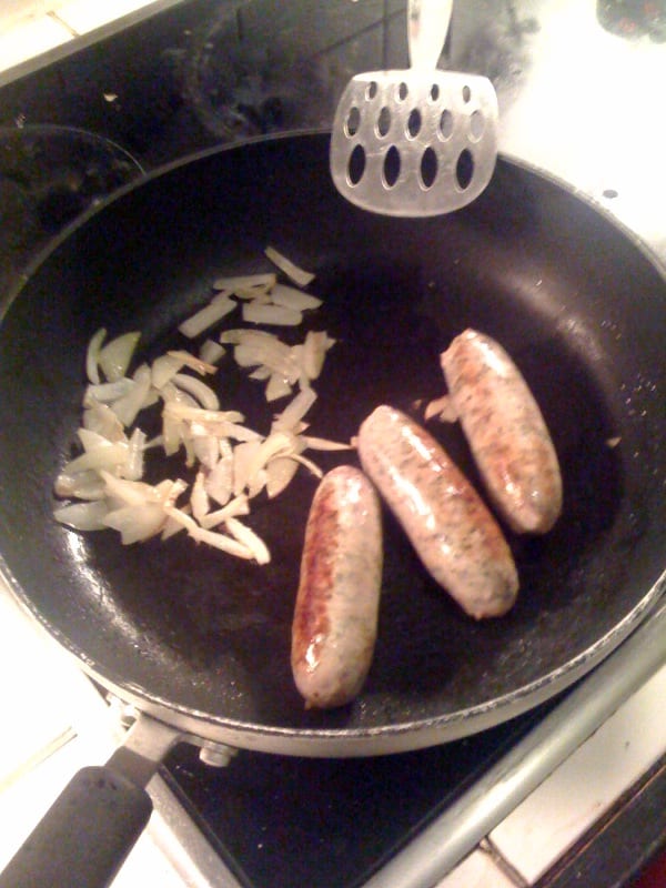 Two thick Lincolnshire sausages browning whole in a pan — the coarse, sage-forward everyday link