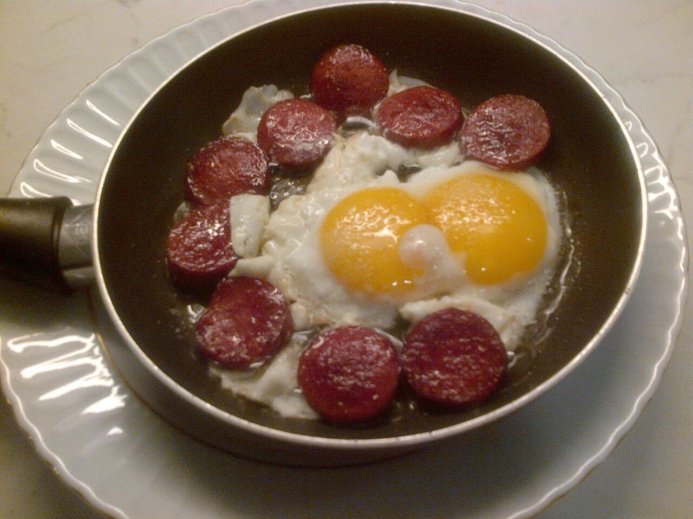 Slices of sucuk curling and blistering in a dry pan, rendering their own orange fat, one egg cooking beside them